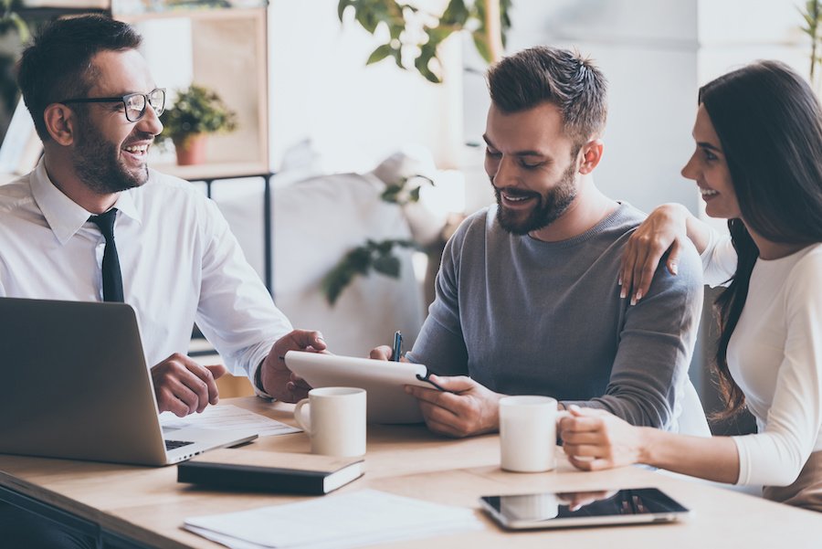 Just put your signature here! Cheerful young man signing some documents while sitting together with his wife and man in shirt and tie